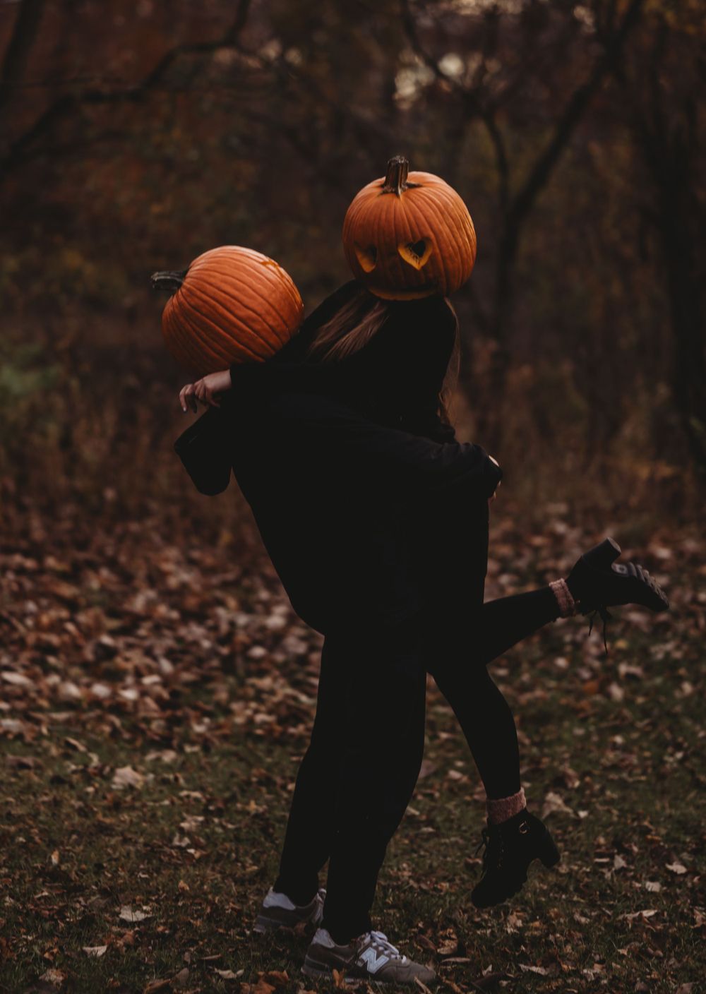 Person in black clothing holding carved pumpkins while posing in fallen leaves.