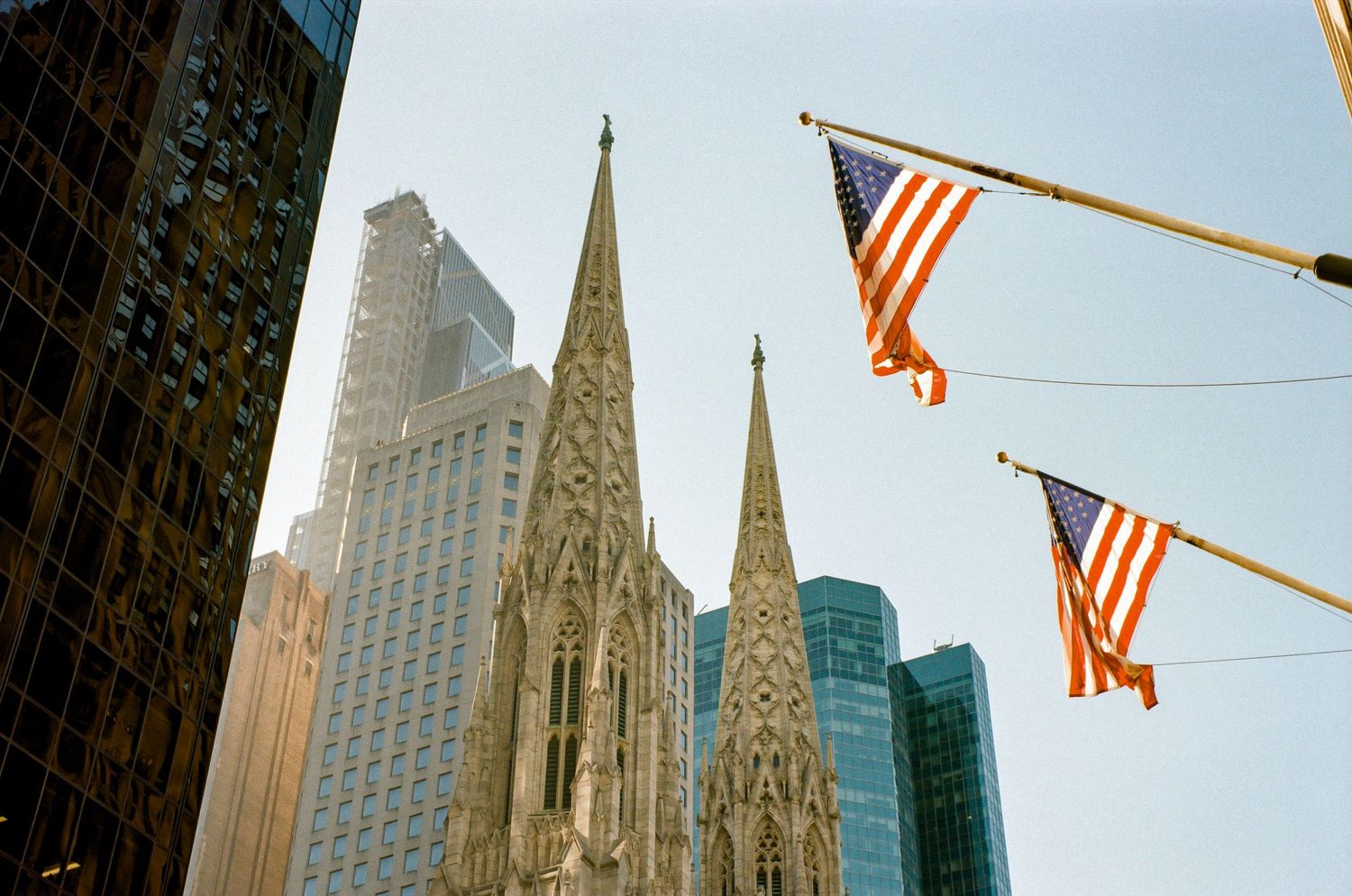 Gothic spires of St Patrick's Cathedral rise against Manhattan skyscrapers with American flags waving above.