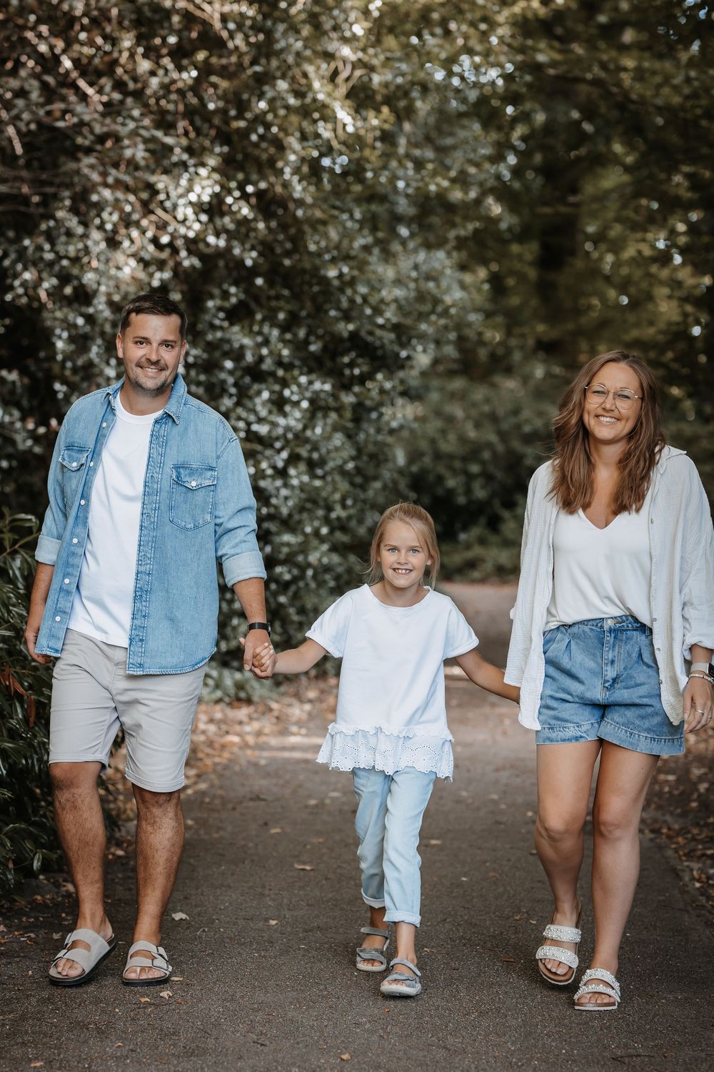 Family of three walking together on tree-lined path in casual summer outfits.