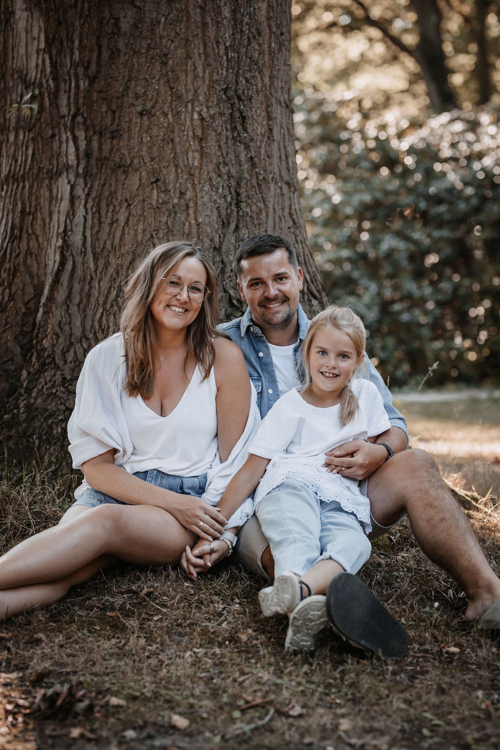 Family of three posing together against large tree trunk in forest setting.