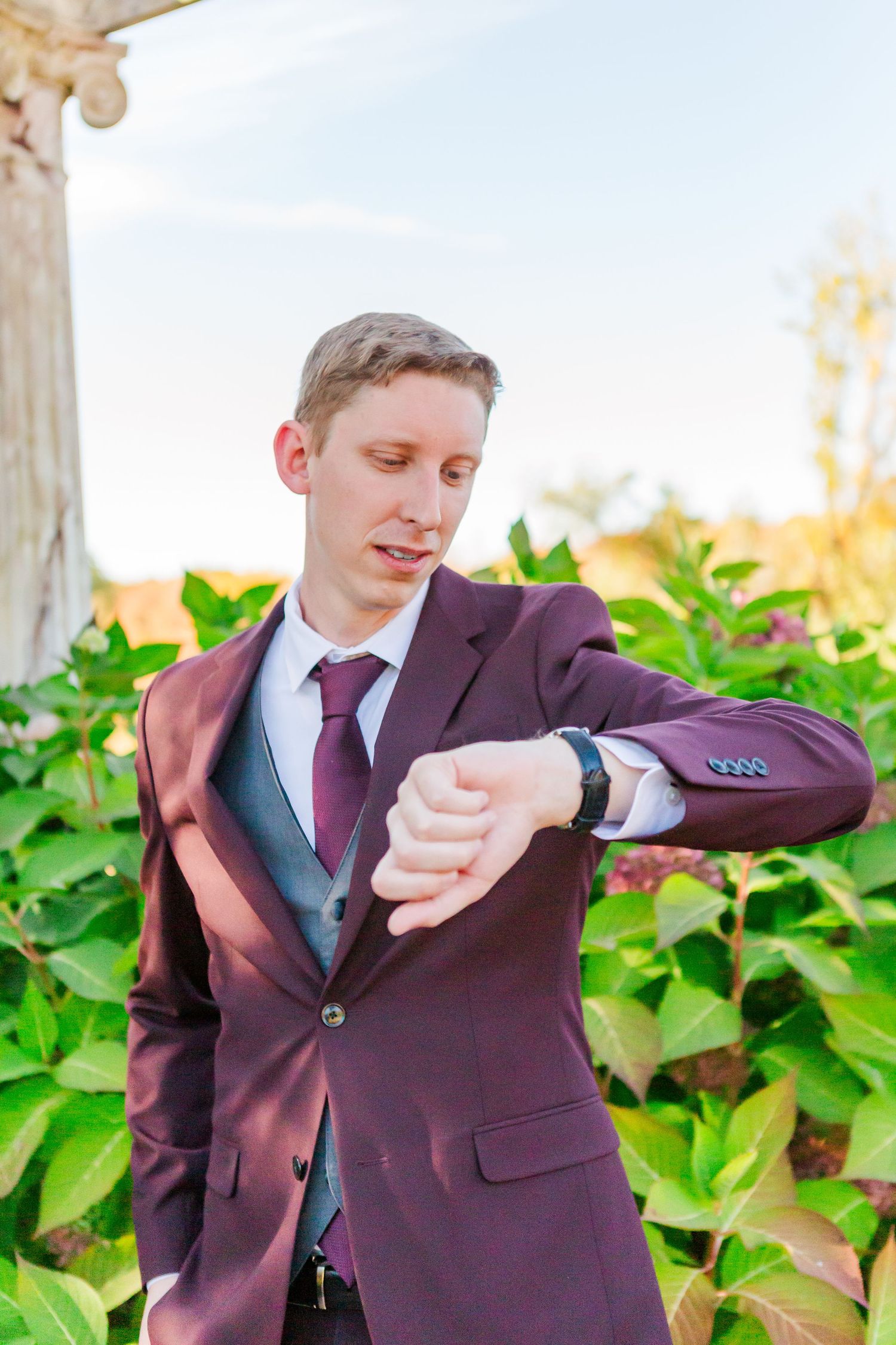 Groom in burgundy suit checking time on wristwatch in garden setting.