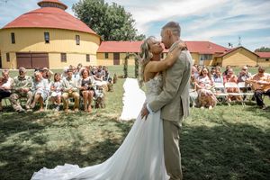 A Couple shares an intimate outdoor kiss during their wedding ceremony at a rustic farm venue with guests watching.