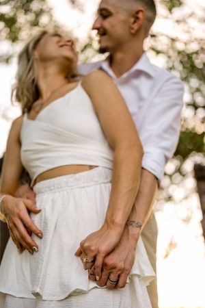 A romantic couple in white outfits holding hands and laughing together in soft natural light outdoors.