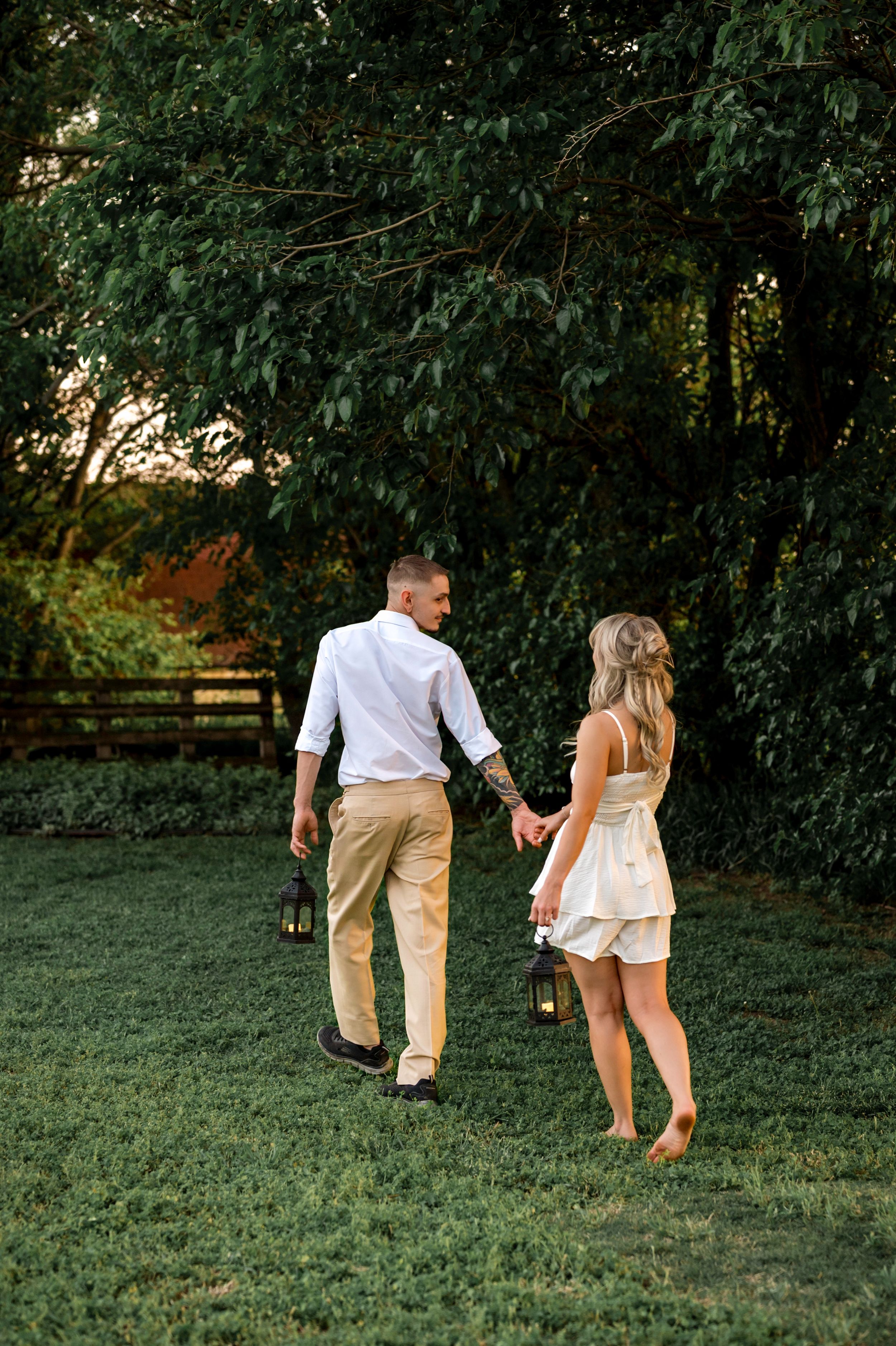 Couple walking away through a lush green lawn with tall trees in the background during a sunny summer day.