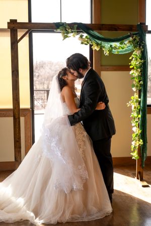 A romantic wedding moment between newlyweds under a green and floral arch by a window.