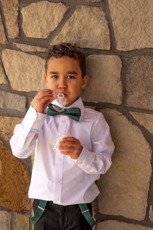 A well-dressed child wearing a white shirt with bow tie enjoys a snack against a stone wall.