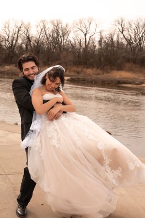 A couple shares a romantic embrace in formal wedding attire by a river with bare winter trees in the background.