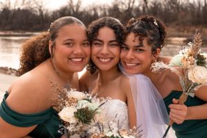 Three close friends share a joyful moment at a wedding celebration, holding bouquets against a natural backdrop.