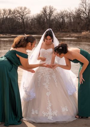 A wedding party in emerald green dresses helps adjust a bride's flowing lace gown by a serene waterfront setting.