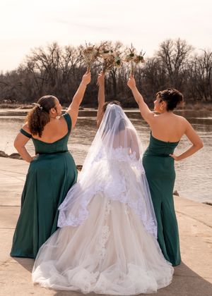 Three women in emerald green dresses stand at a waterfront, raising their bouquets in celebration.