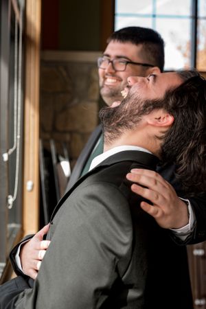 Two people in suits and glasses embrace while laughing joyfully in a warmly lit indoor setting.