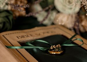 Wedding rings and invitation with dark green ribbon displayed on rustic background with soft floral accents.
