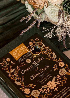 Elegant wedding invitation with gold floral design and wedding rings on dark background with roses and dried flowers.