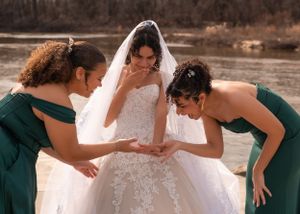 Bridesmaids in emerald green dresses admire a bride's lace wedding gown beside a lake.