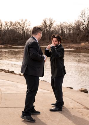 Two people in suits stand by a river during sunset, one proposing to the other in a sepia-toned moment.