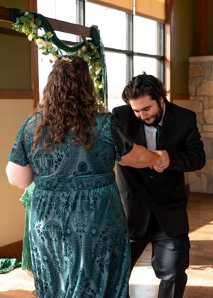A couple performs a traditional dance move in a room decorated with floral garlands and natural window light.