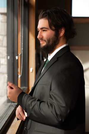 A person with dark hair and beard wearing a black suit and green tie gazes out a window thoughtfully.