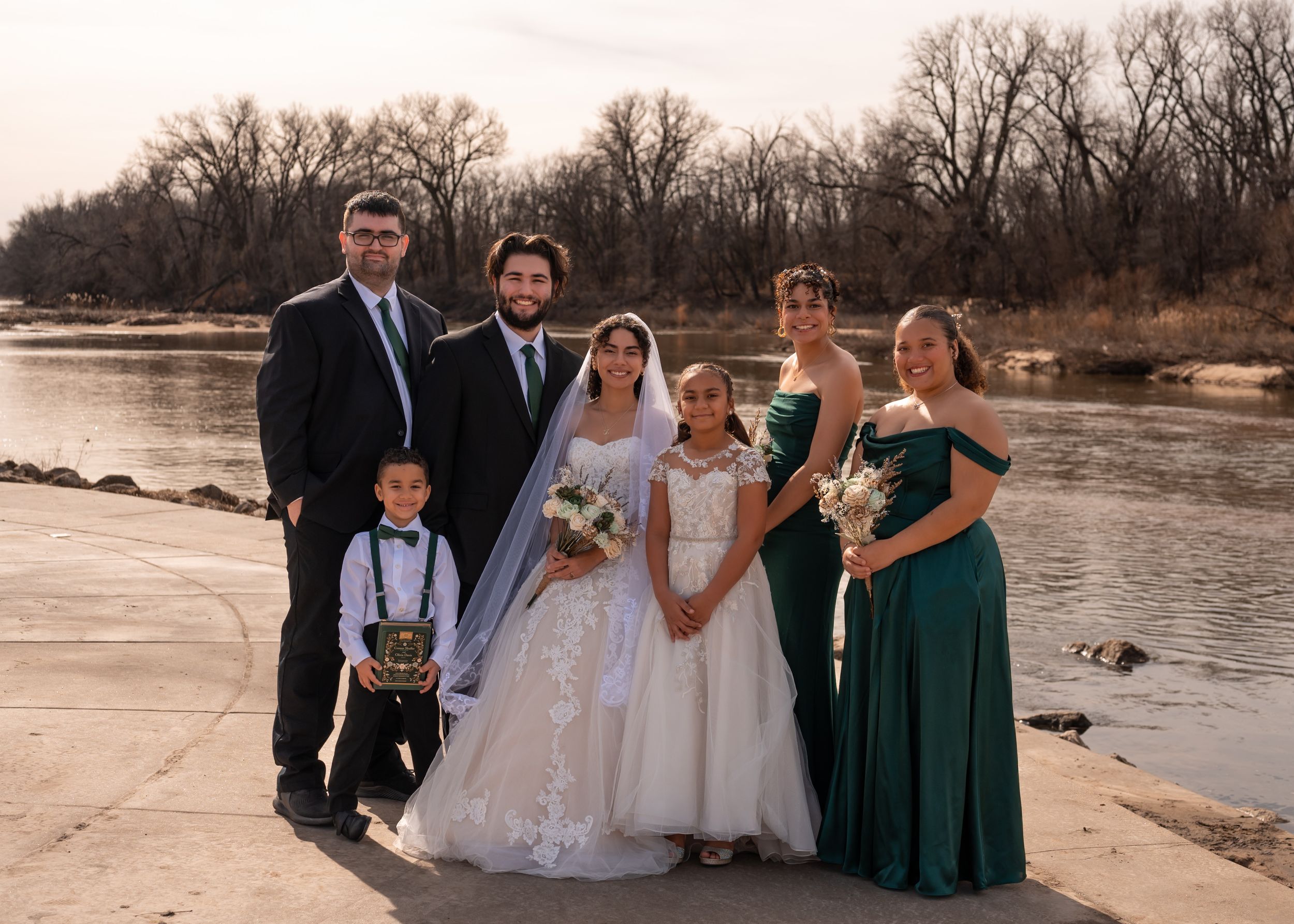 A wedding party poses together on a riverbank at sunset wearing formal attire in navy and emerald green.