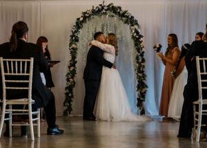 Newlyweds share their first kiss under a floral arch during an indoor wedding ceremony.