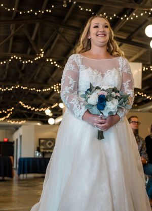 A beautiful bride in a lace-sleeved white gown holds a blue and white floral bouquet under string lights.