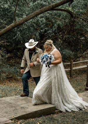 Bride in white gown walks with companion in cowboy hat along forest path.