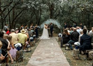 Outdoor wedding ceremony with guests seated on white chairs under tree canopy.