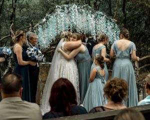 Wedding party gathers under light blue floral arch during outdoor ceremony.