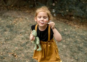 A joyful child in mustard yellow overalls blows bubbles while playing outdoors.