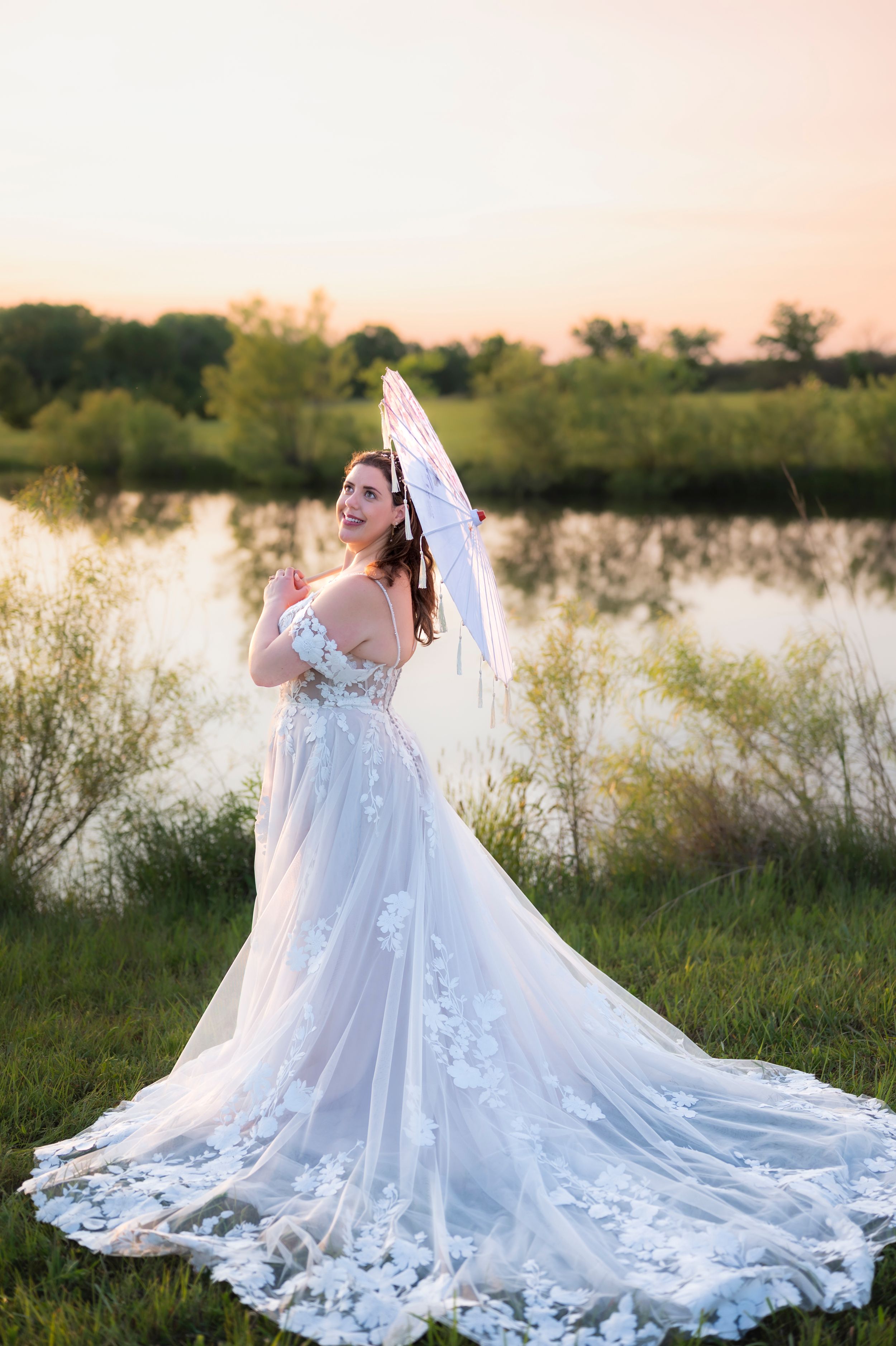 A bride in a flowing white lace gown with long train poses at sunset by a serene lake with trees in the background.