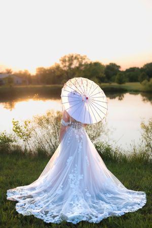 A bride in a flowing lace wedding dress holds a white parasol while standing by a tranquil lake at sunset.