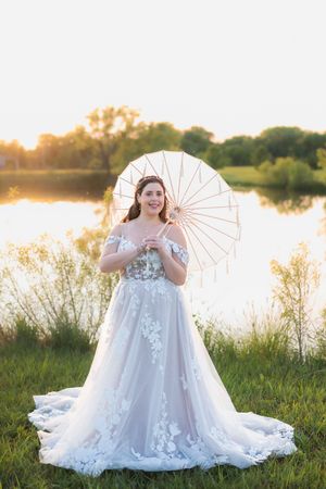 A bride in a flowing white lace dress poses with a parasol by a serene lake at golden hour.