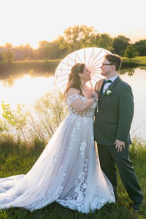 A romantic wedding couple poses by a tranquil lake at sunset, with a white parasol and a flowing lace gown.