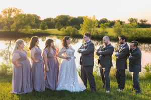 Wedding party in purple dresses and dark suits pose together at sunset by a scenic lake.