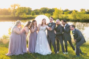 Bridal party laughs together while posing outdoors near a lake at golden hour.