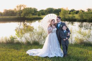 Wedding photo by a serene lake at sunset with a white parasol and natural landscape in the background.