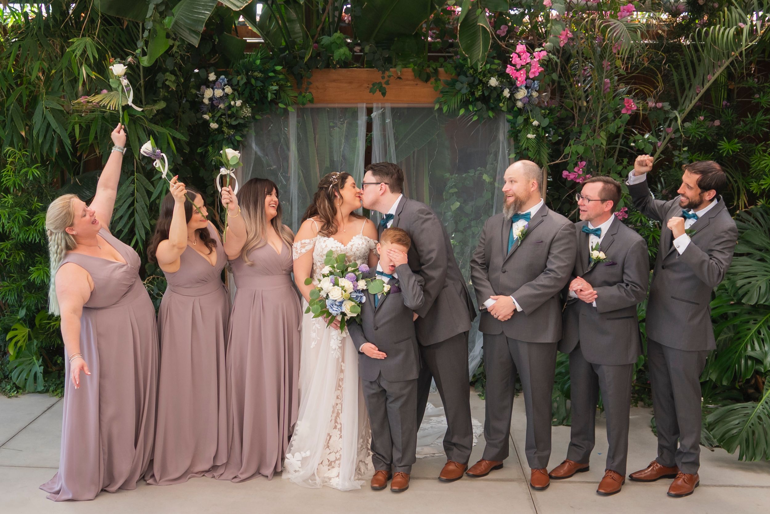 Wedding party members in gray suits and mauve dresses pose in line for formal photos outdoors with tropical greenery.