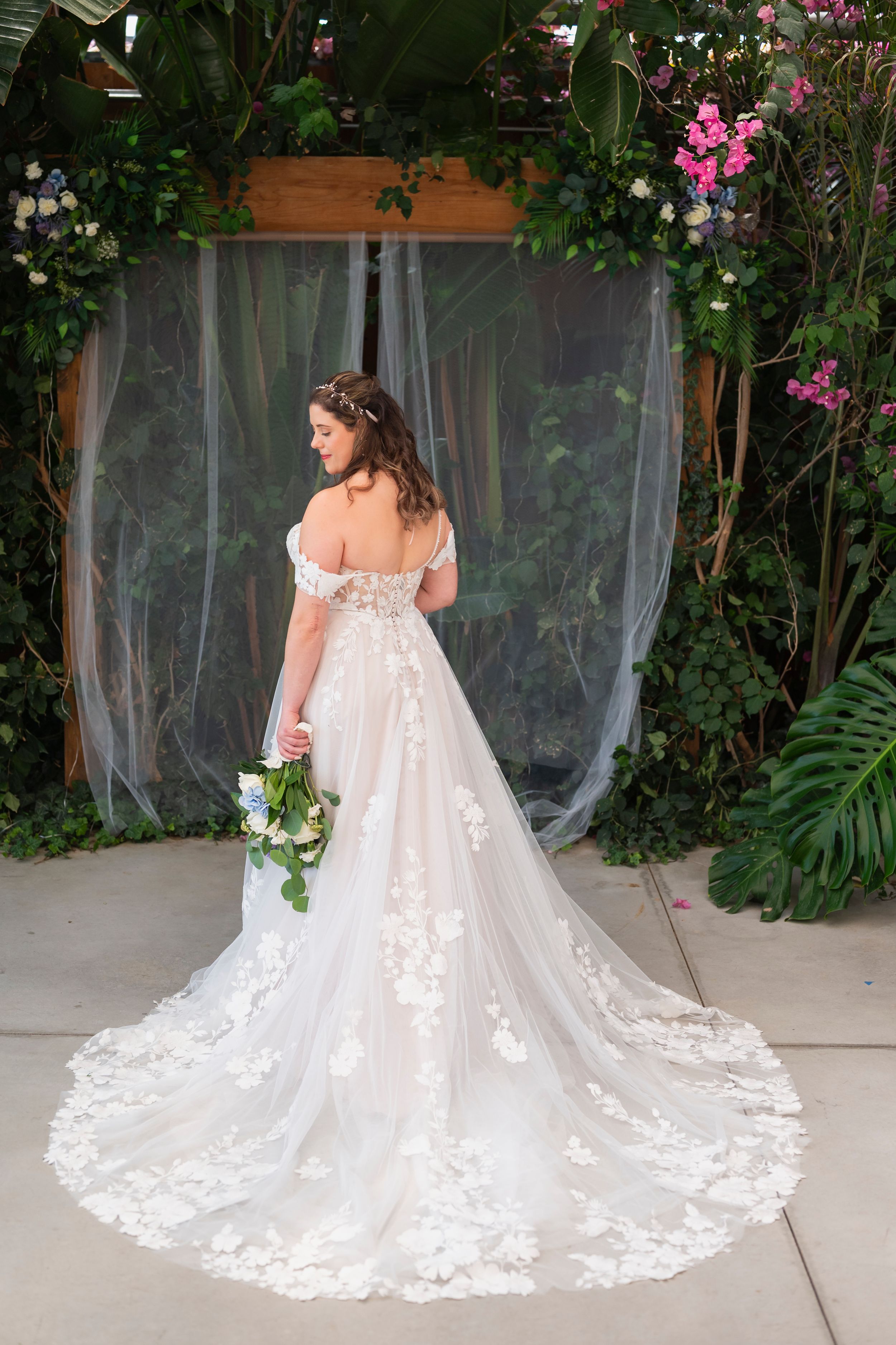 Back view of a bride in an off-shoulder lace wedding gown with long train standing against a floral garden backdrop.