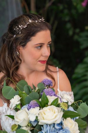 A bride wearing a delicate crystal hairpiece holds a bouquet with white roses, blue hydrangeas and purple flowers.
