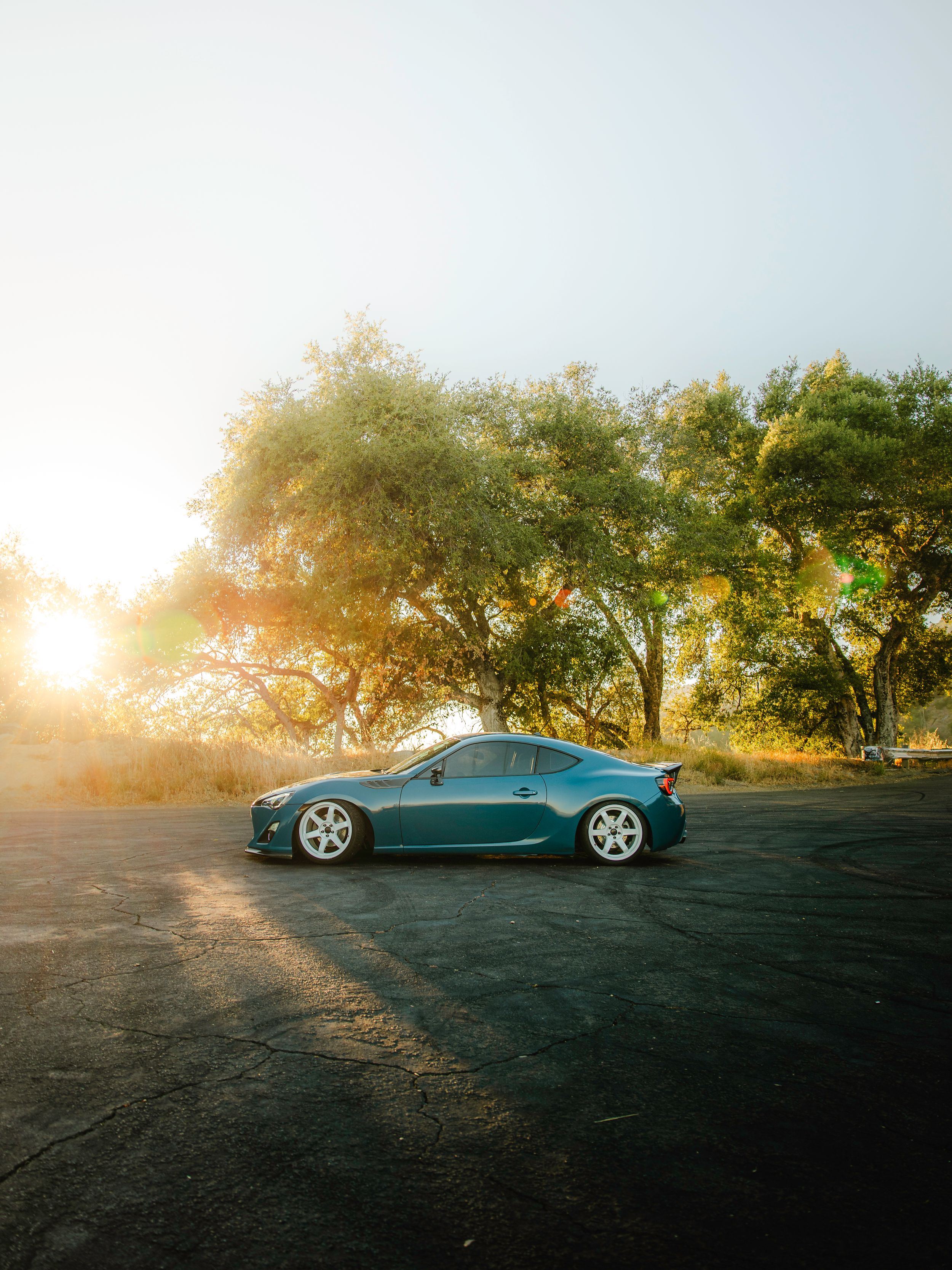 A teal Toyota GT86 parked on asphalt with sunlit trees in the background during golden hour.