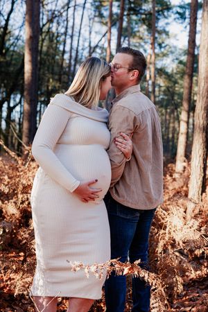 Couple embraces in a romantic forest setting with golden sunlight filtering through tall pine trees.