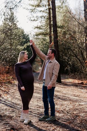 A couple dances together on a forest path during autumn, surrounded by tall trees and fallen leaves.