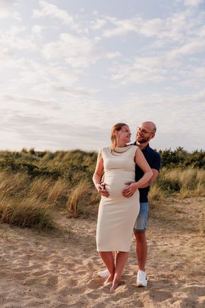 A couple embraces on a sandy beach with tall grass and cloudy skies in the background.