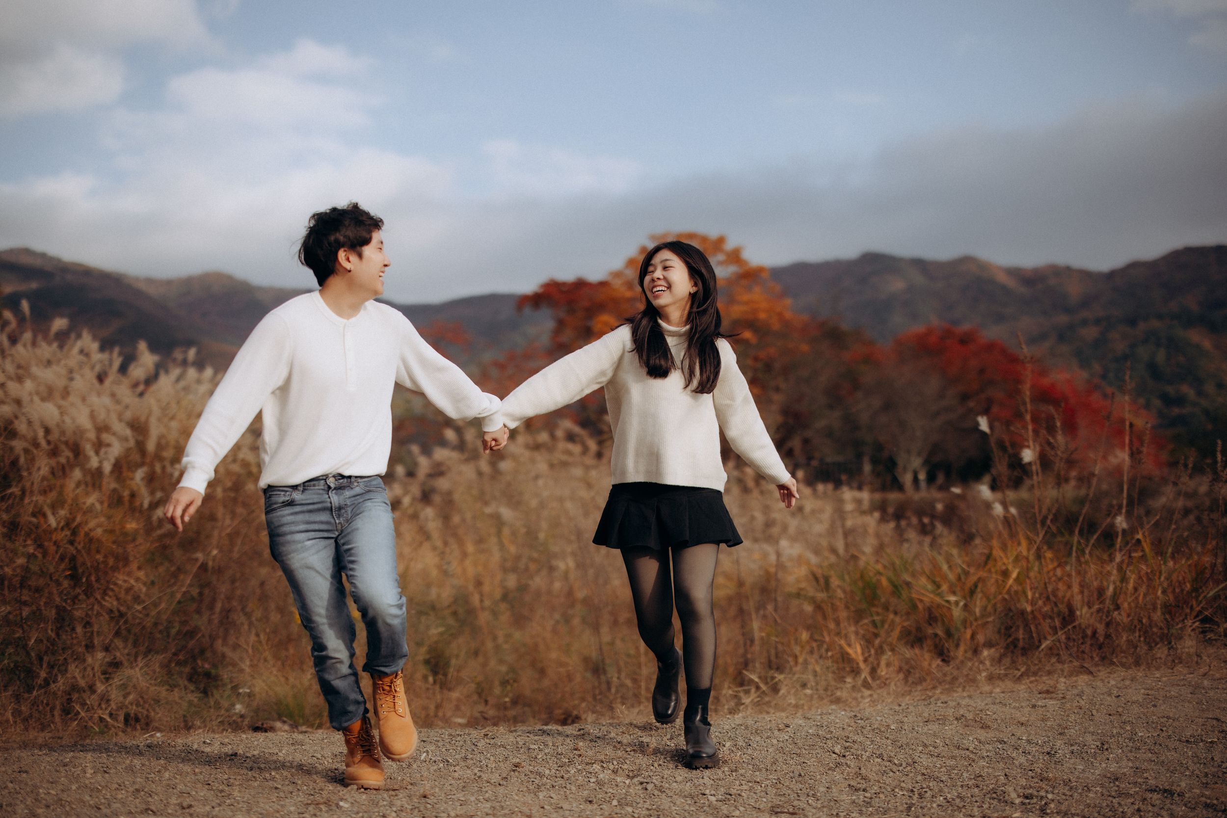 A couple holds hands while walking together at Lake Kawaguchiko region during autumn with red foliage in background.