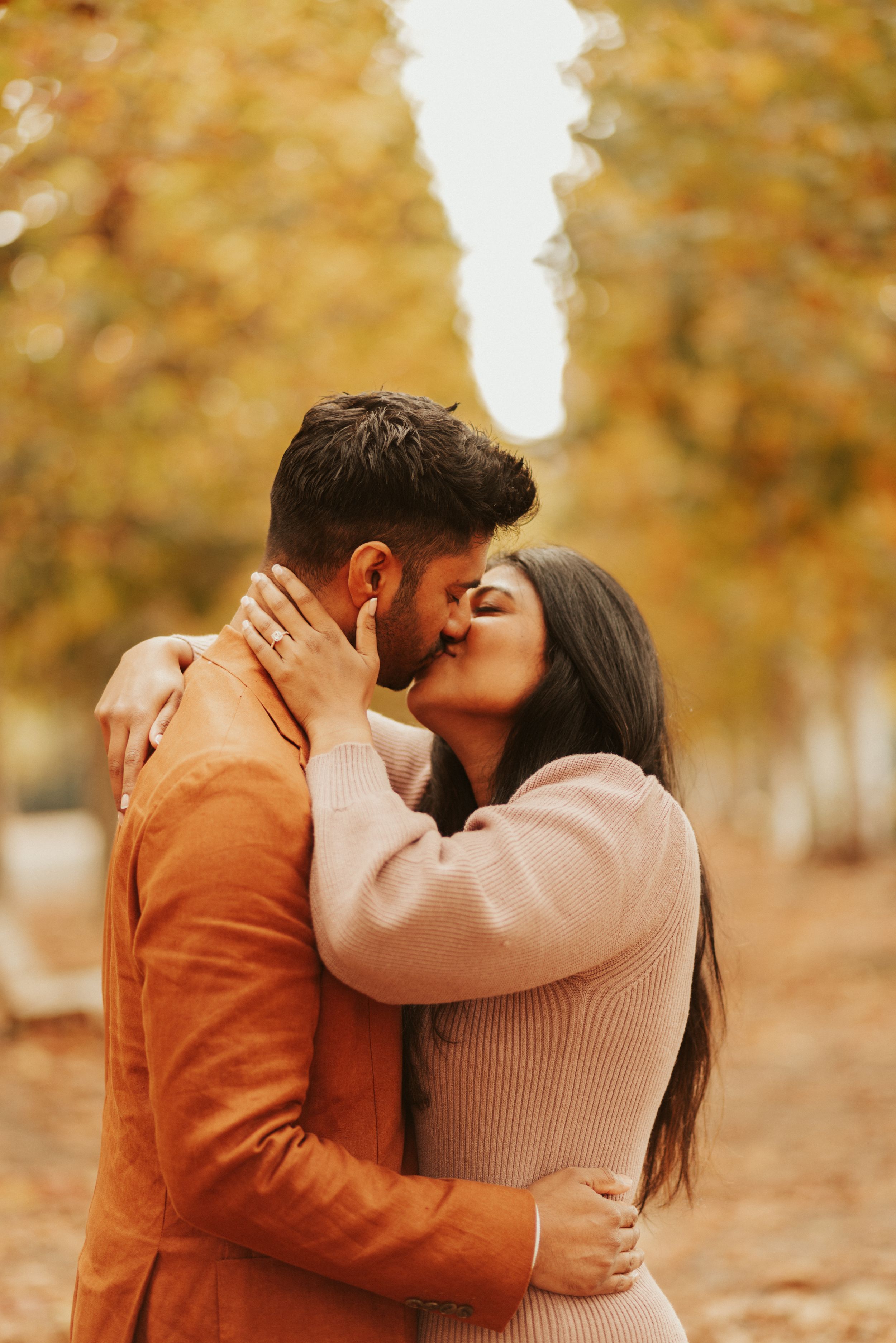 A romantic couple share a tender kiss while embracing in an autumn park with golden foliage in the background.