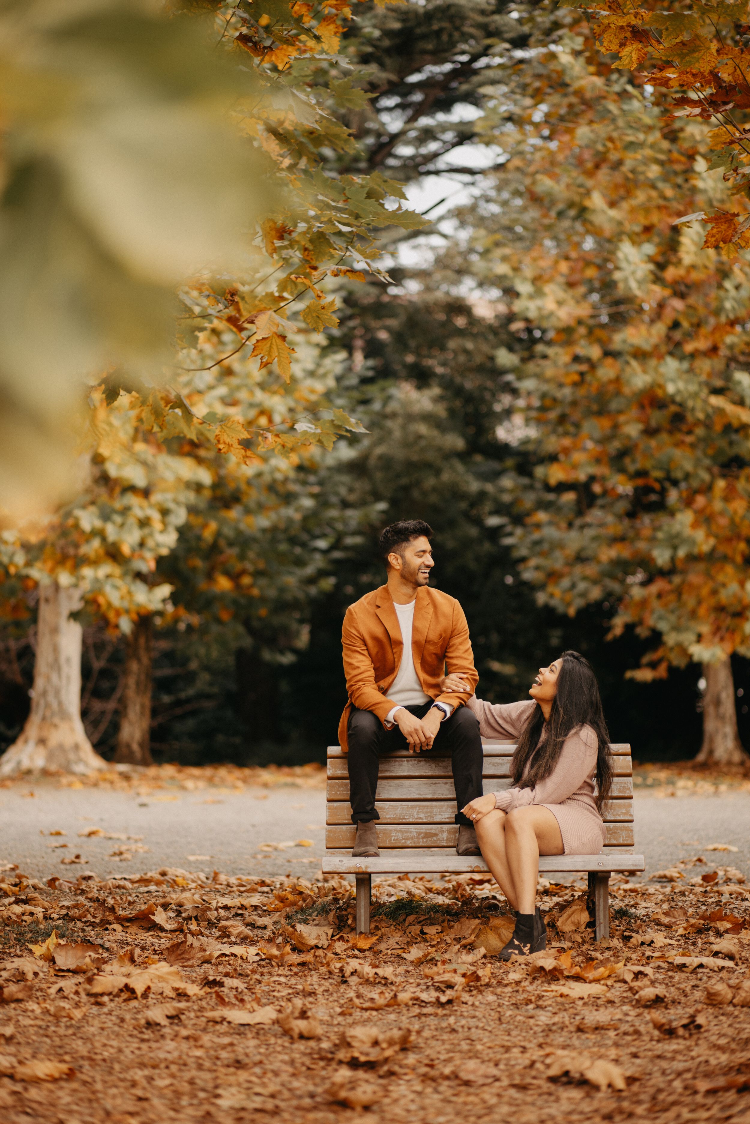 Two people sit on a park bench surrounded by autumn leaves and orange foliage in a romantic fall setting.