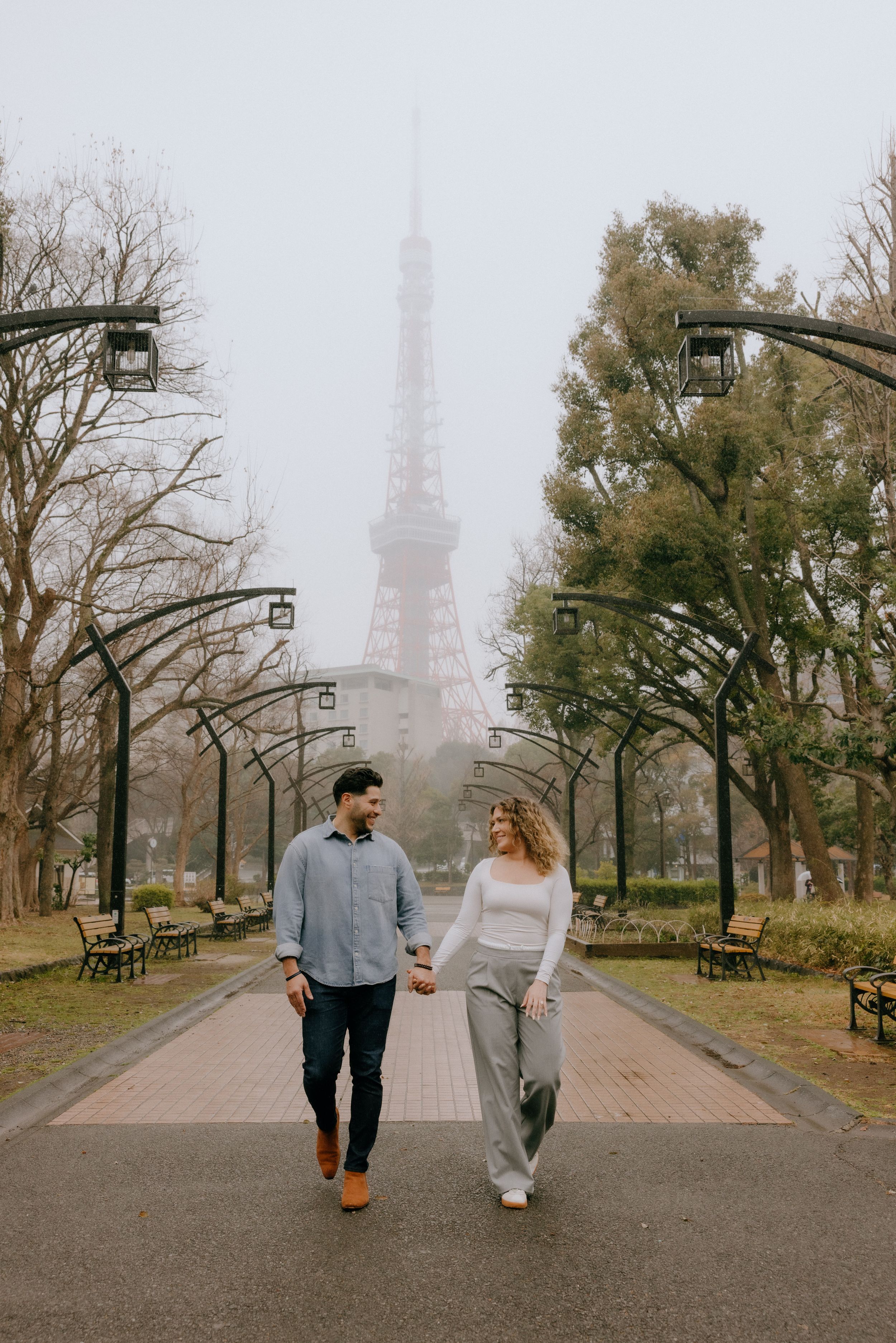 A romantic couple walking hand in hand down a foggy path with the Eiffel Tower visible in the misty background.