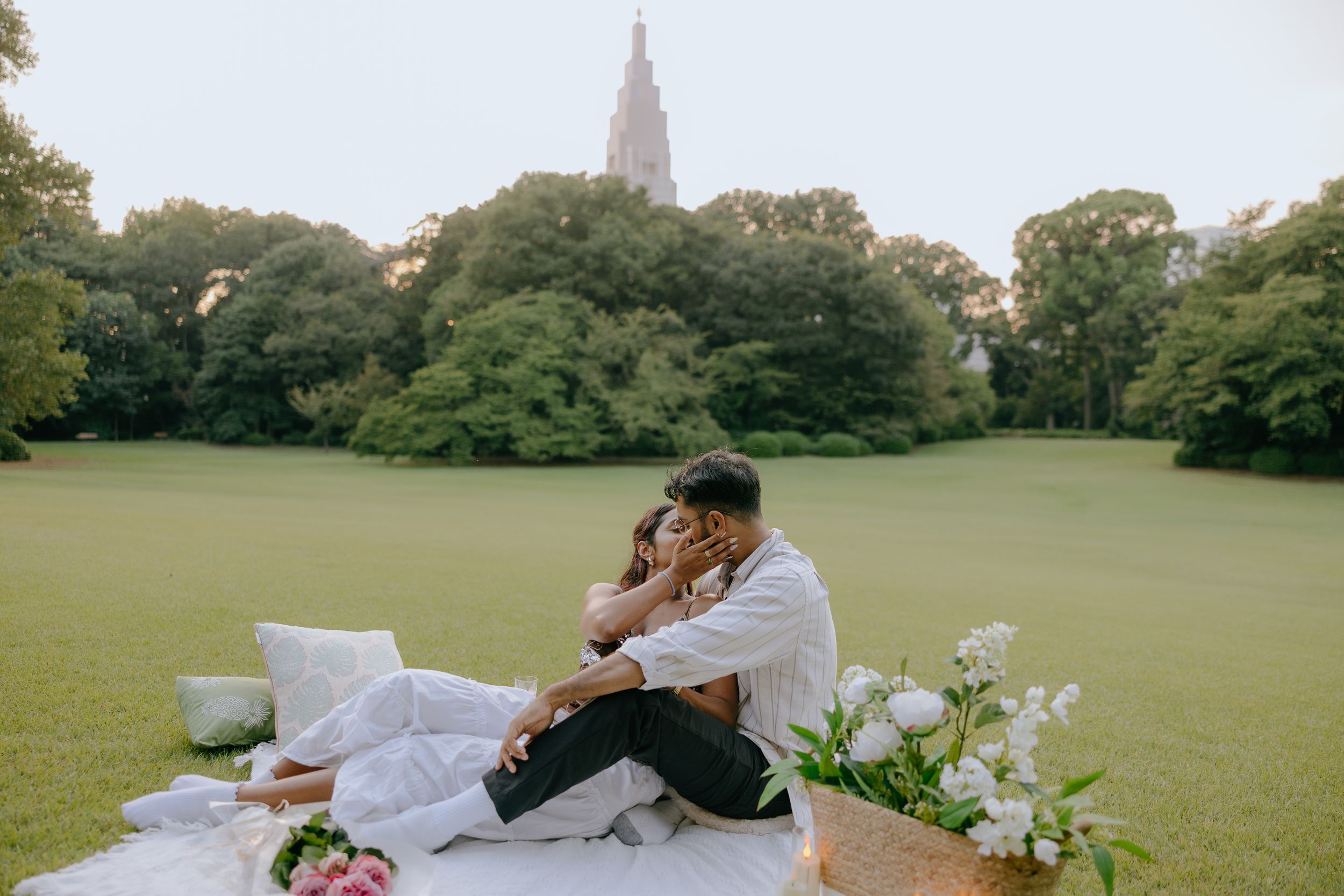A couple enjoys a romantic picnic with wine and flowers in a lush green park during golden hour.