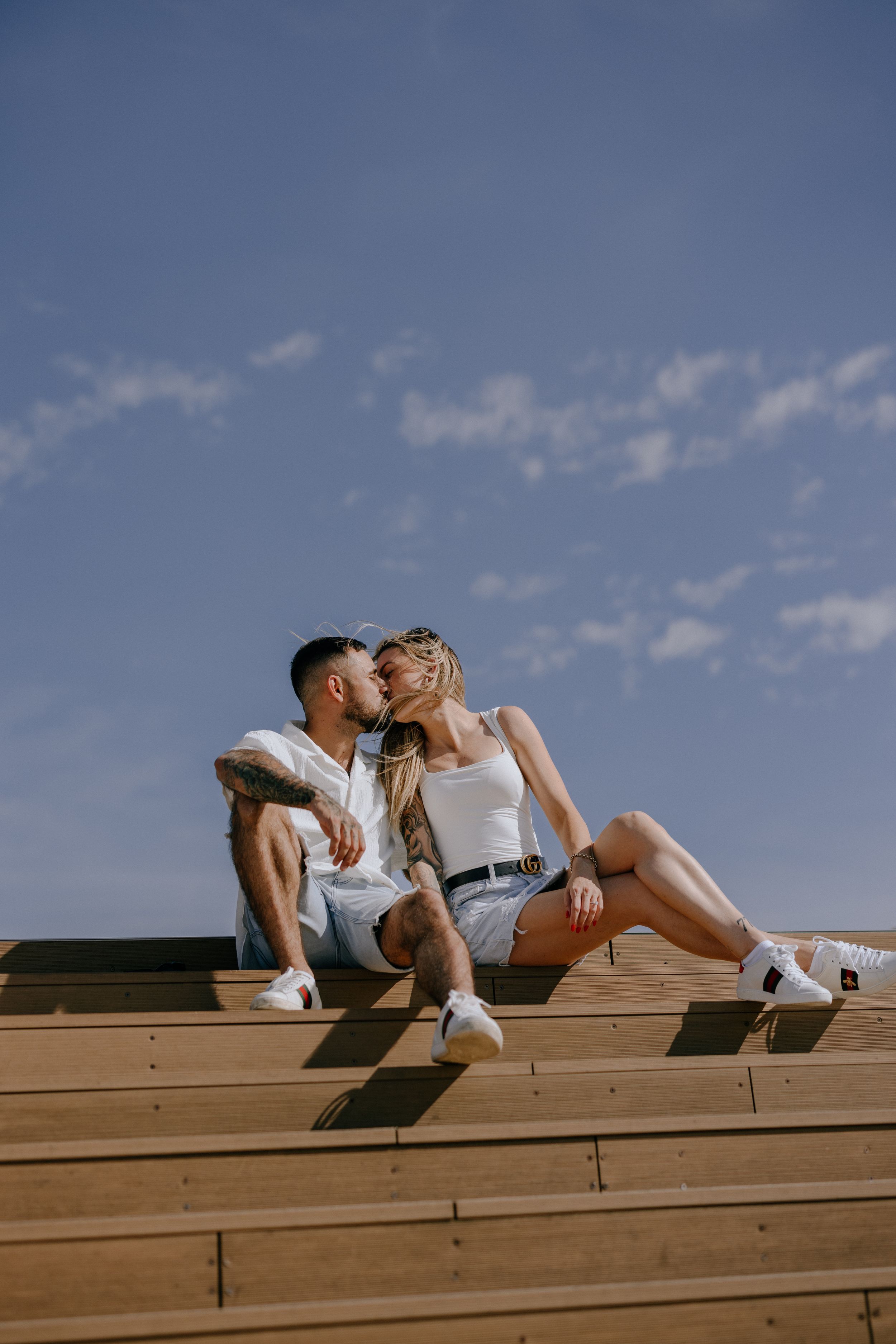 A casual couple sharing an intimate moment on wooden steps against blue sky.
