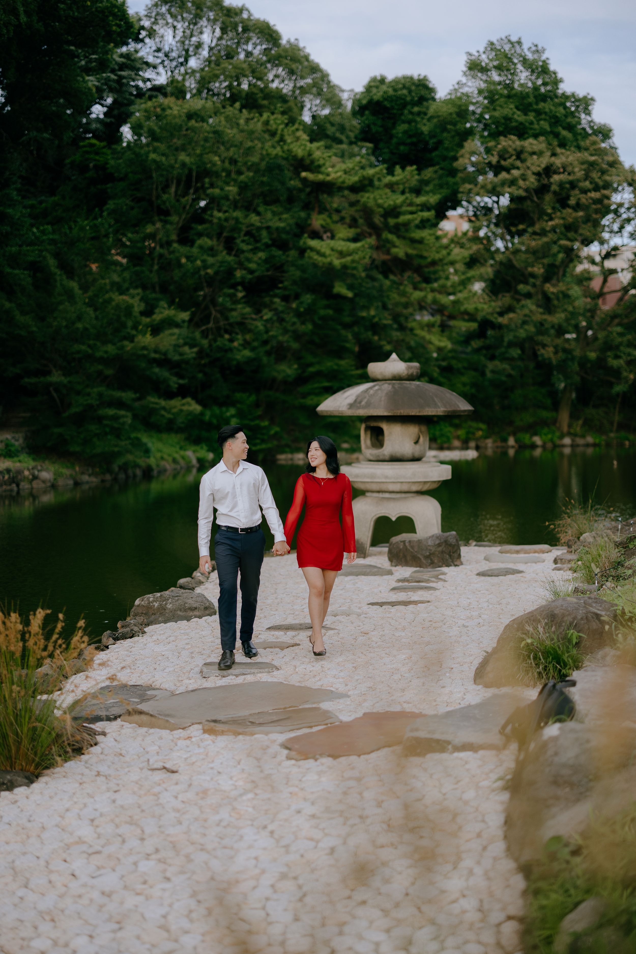 A couple walks together on a winding stone path near a serene Japanese garden pond with a traditional stone lantern.