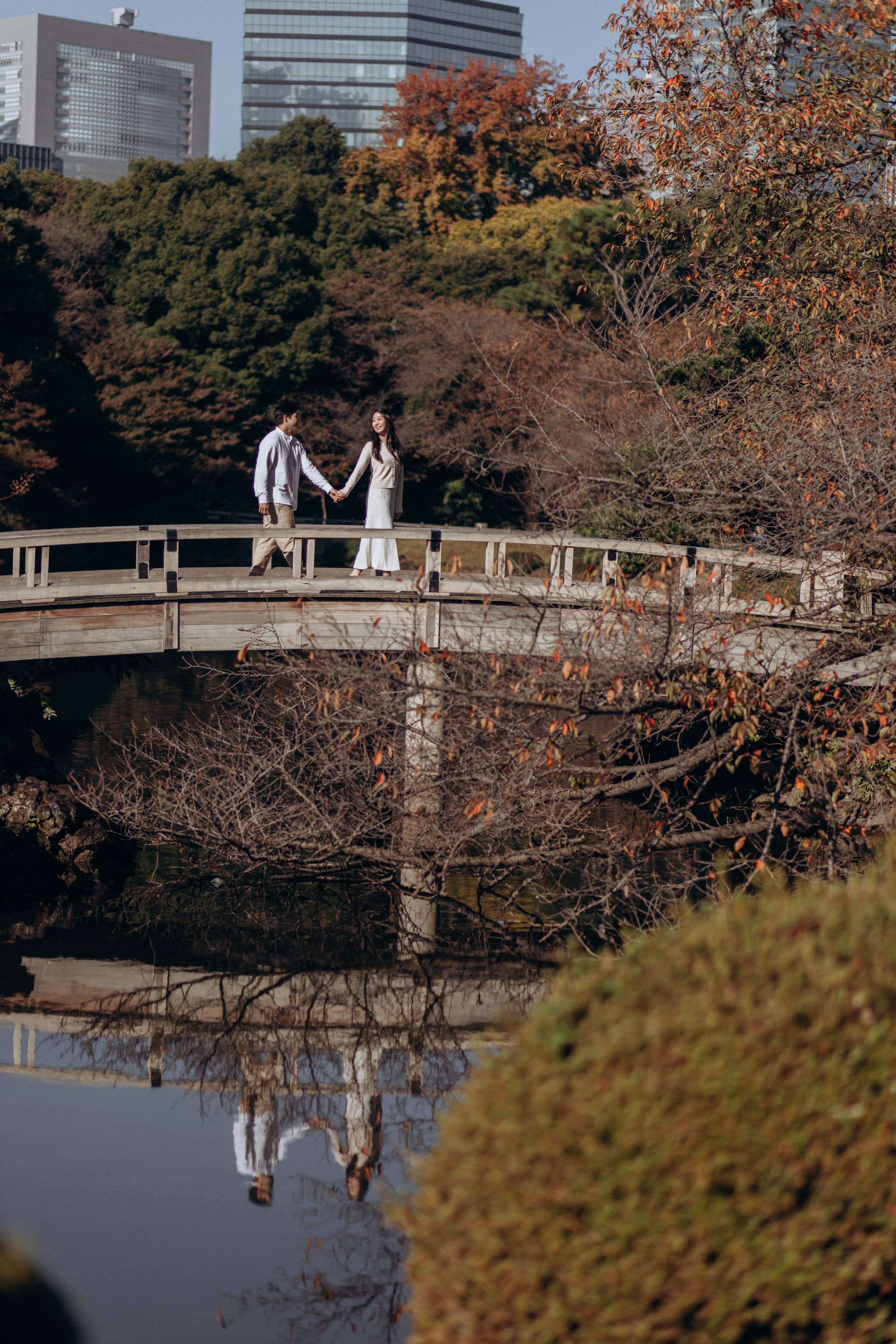 Two people stand on a wooden bridge with autumn foliage and modern buildings in the background at a Japanese garden.